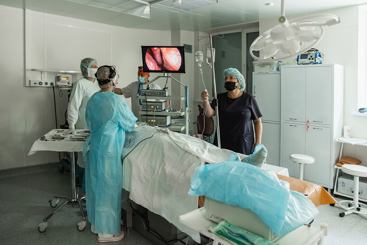 A surgical team works together in a clean, modern operating room. They monitor a patient on the table, using monitors, IVs, and sterile equipment during a live procedure.